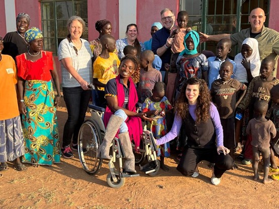 Purple Vest delegation members from Access Israel pose with local children and adults, including a smiling woman in a wheelchair, during an inclusive visit in Uganda.